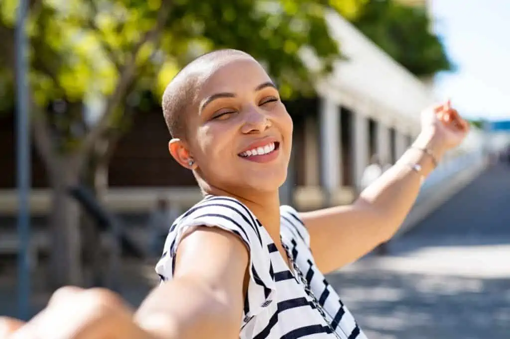 Cheerful young bald woman spreading arms feeling happy. Smiling girl feeling free with closed eyes outdoor. Happy curvy woman enjoying the day while on holiday.