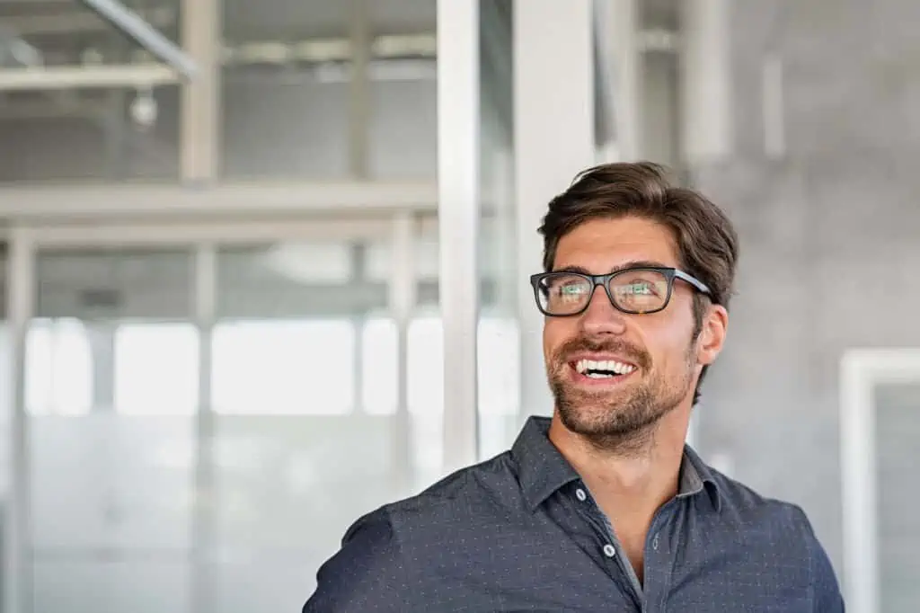Confident young businessman looking up with big grin. Happy handsome business man wearing spectacles looking away in modern office. Close up face of smiling guy having idea. Man dreaming of success and pondering new startup.