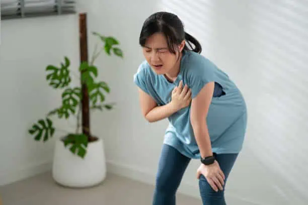 Asian woman suffering from chest pain breathing problems while exercising in living room at home.